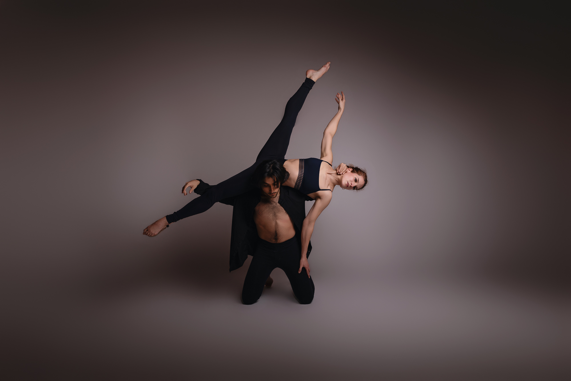 Séance photo en studio à Maringues: Lucile Duvert Photographe. Deux danseurs contemporains habillés en noir réalisent une figure de portée. L'homme est agenouillé au sol et soutient la danseuse qui s'étire en arabesque, une jambe et un bras levés vers le ciel, dans une pose expressive et équilibrée. Fond neutre aux tons chauds et sombres.