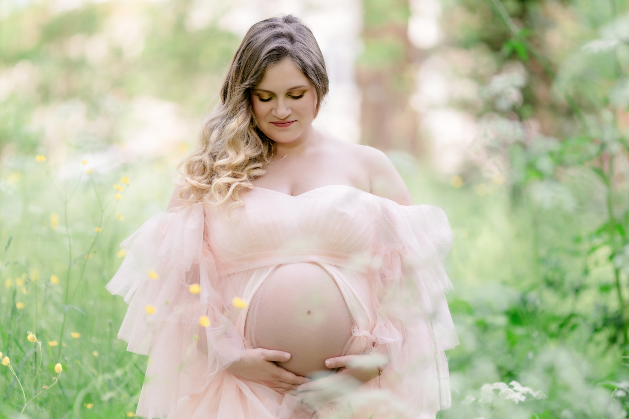 Femme enceinte en robe rose pâle vaporeuse, posant dans une prairie fleurie au parc des Bourins à Vichy. Lumière naturelle douce et ambiance poétique. Séance photo grossesse par Lucile Duvert Photographe